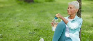 Mature woman wearing sports outfit sitting relaxed on mat in park opening bottle with fresh water