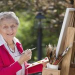 A smiling senior woman is in an outdoor setting painting. Horizontal shot.